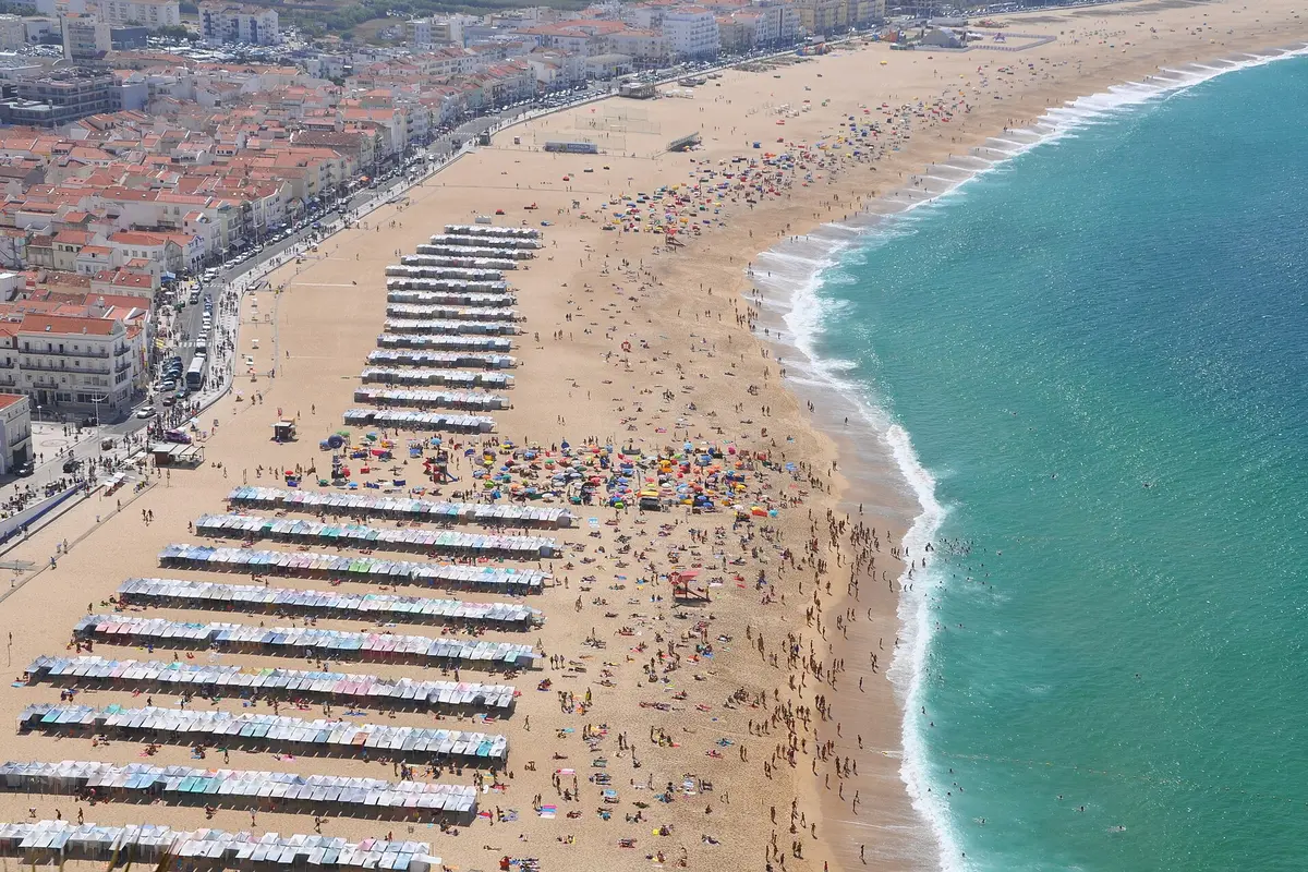 The long town beach of Nazaré seen from above, with rows of beach tents, red-roofed houses on the left and surf breaking on the right