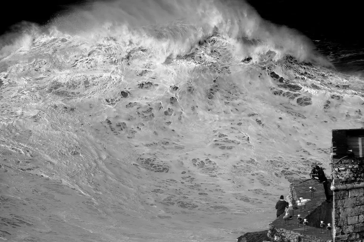 A huge breaking wave at Praia do Norte in black and white, with two people on a stone terrace and a stone wall at the lower right for scale